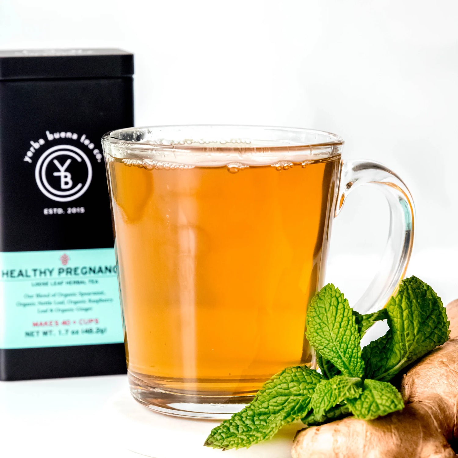 Brewed amber tea in clear glass mug with fresh mint leaves and ginger root beside black canister labeled Healthy Pregnancy herbal tea, by Yerba Buena Tea Company.