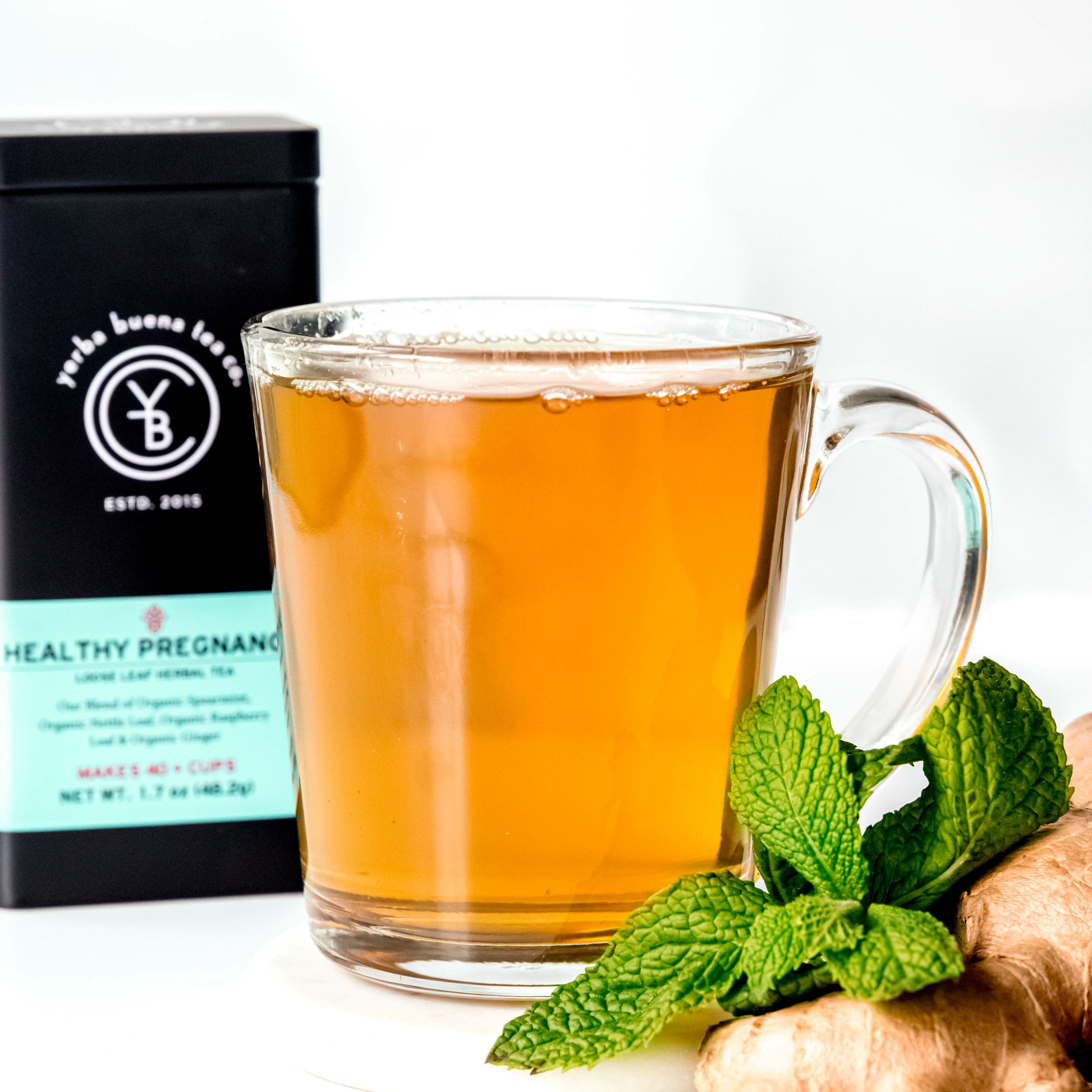 A clear glass cup of Healthy Pregnancy tea, brewed to a golden color, placed beside fresh mint leaves and ginger root, with a Yerba Buena Tea Co. tin in the background.