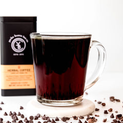 Dark brewed herbal coffee in a clear glass mug surrounded by scattered coffee beans and a black pouch, by Yerba Buena Tea Company.