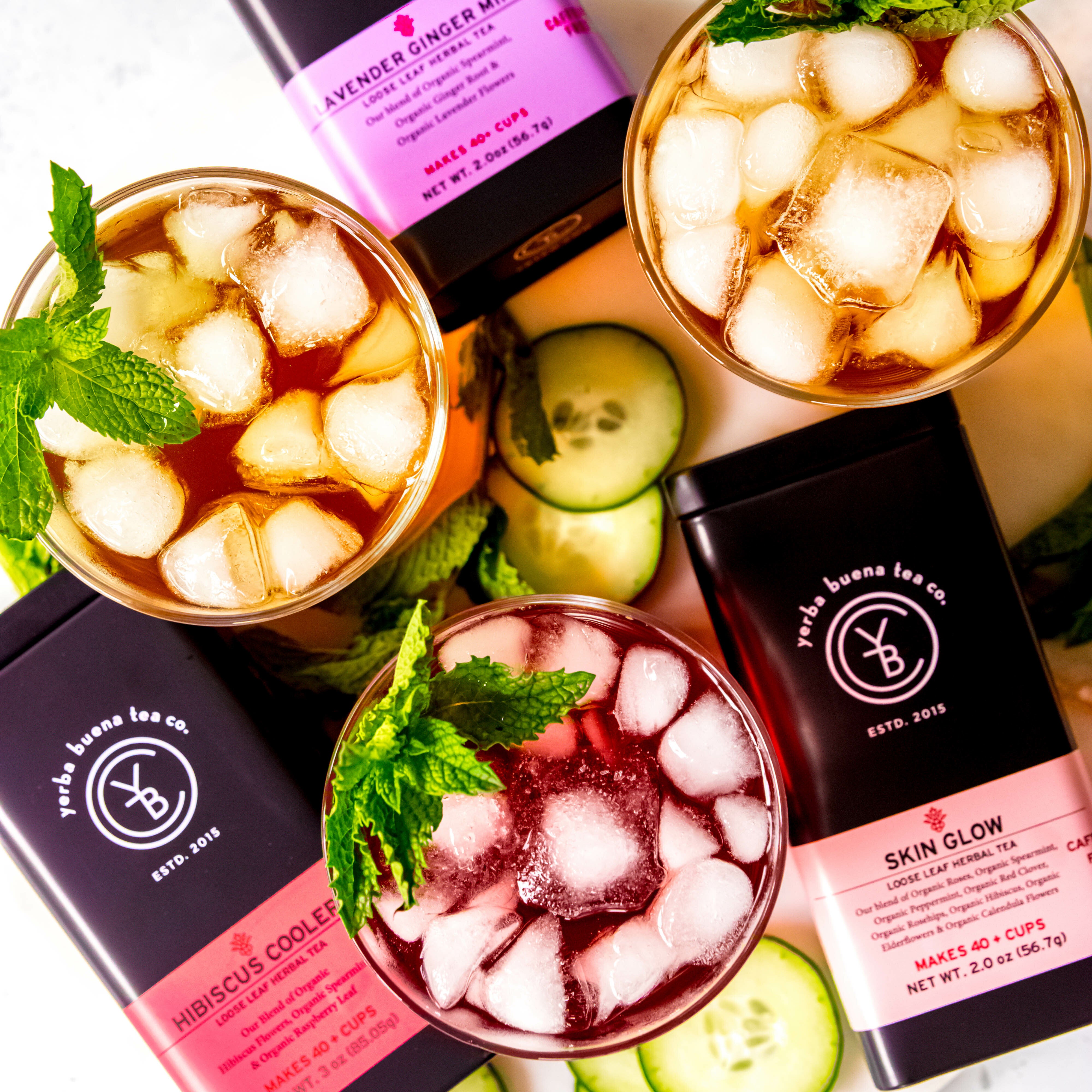 Three glasses of iced tea with fresh mint leaves from Yerba Buena Tea Co., surrounded by tins of Lavender Ginger Mint, Skin Glow, and Hibiscus Cooler teas, along with cucumber slices