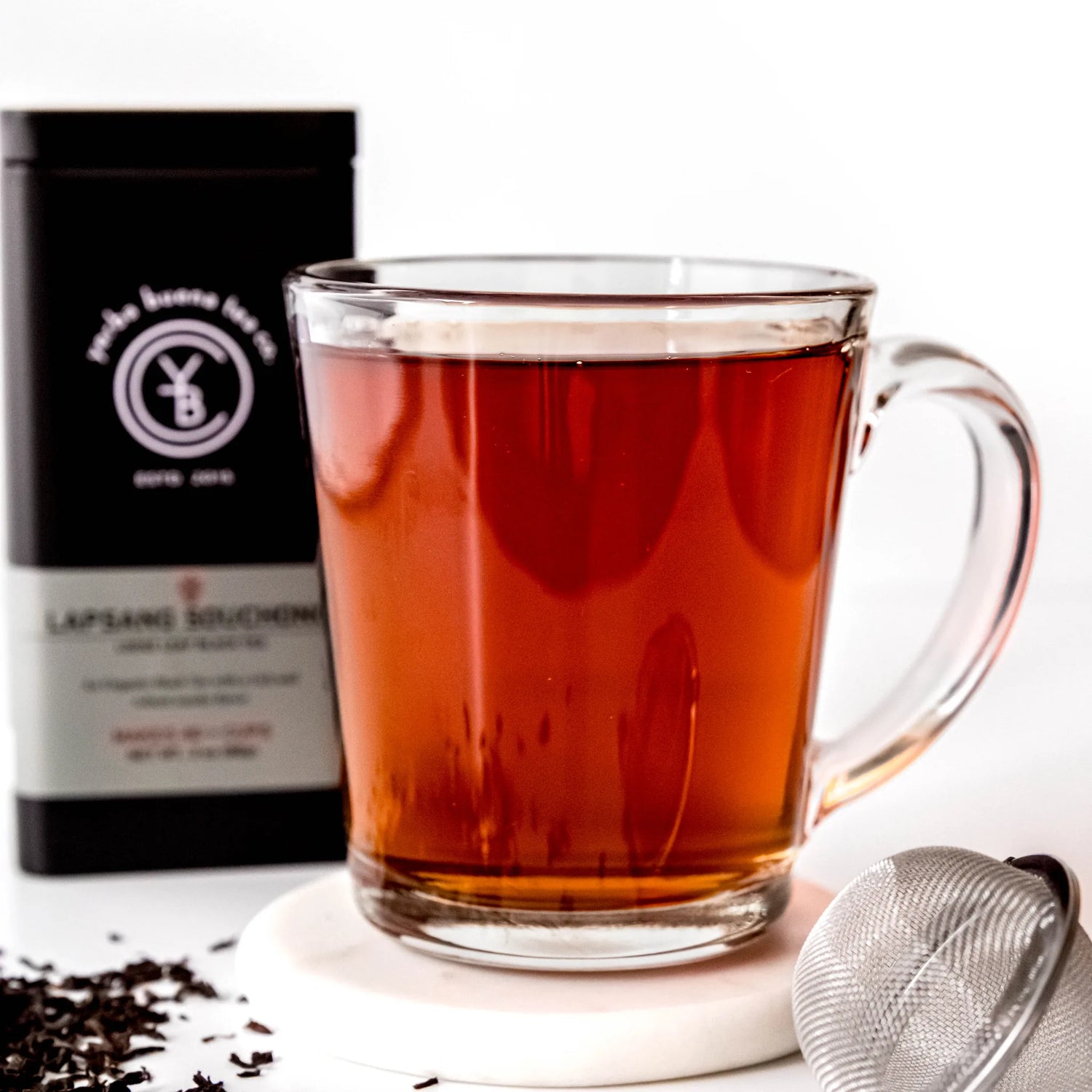 Brewed Lapsang Souchong black tea in glass mug with loose leaf scattered nearby and black pouch packaging, by Yerba Buena Tea Company.
