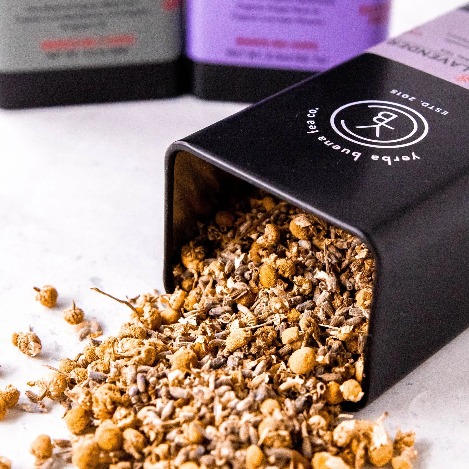 Loose leaf tea spilling from a black pouch labeled Lavender Grey, with purple and white gift boxes blurred in the background, by Yerba Buena Tea Company.
