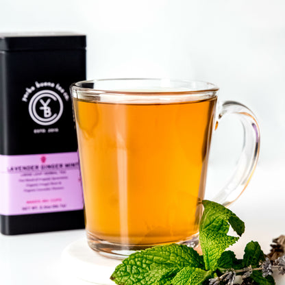 A clear glass cup filled with golden-brown Lavender Ginger Mint tea, placed beside fresh mint leaves and dried lavender flowers, with a Yerba Buena Tea Co. tin in the background.