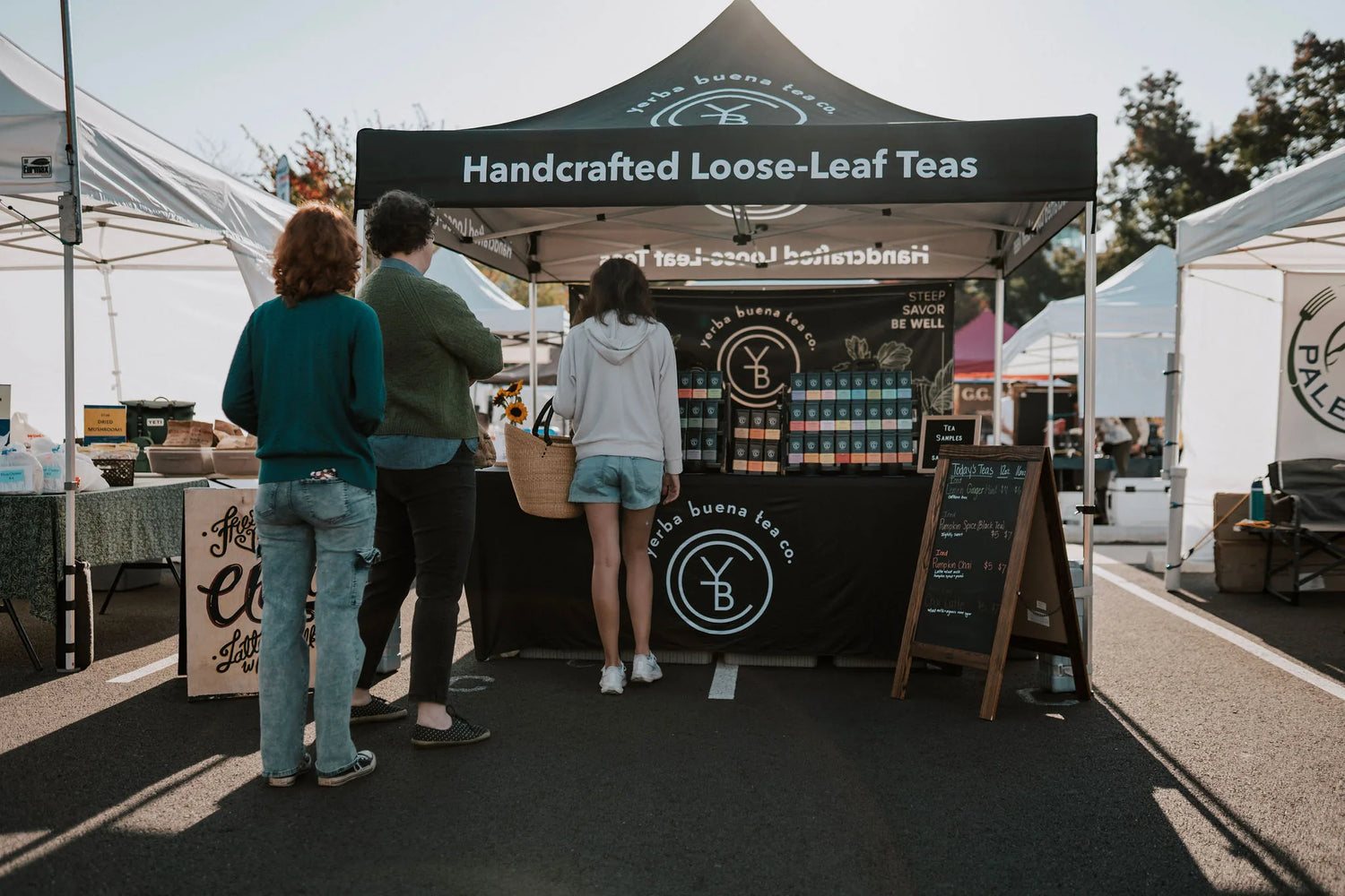 Customers browsing a farmers market booth displaying handcrafted loose-leaf teas with colorful tins, signage, and sampling station, Yerba Buena Tea Company.
