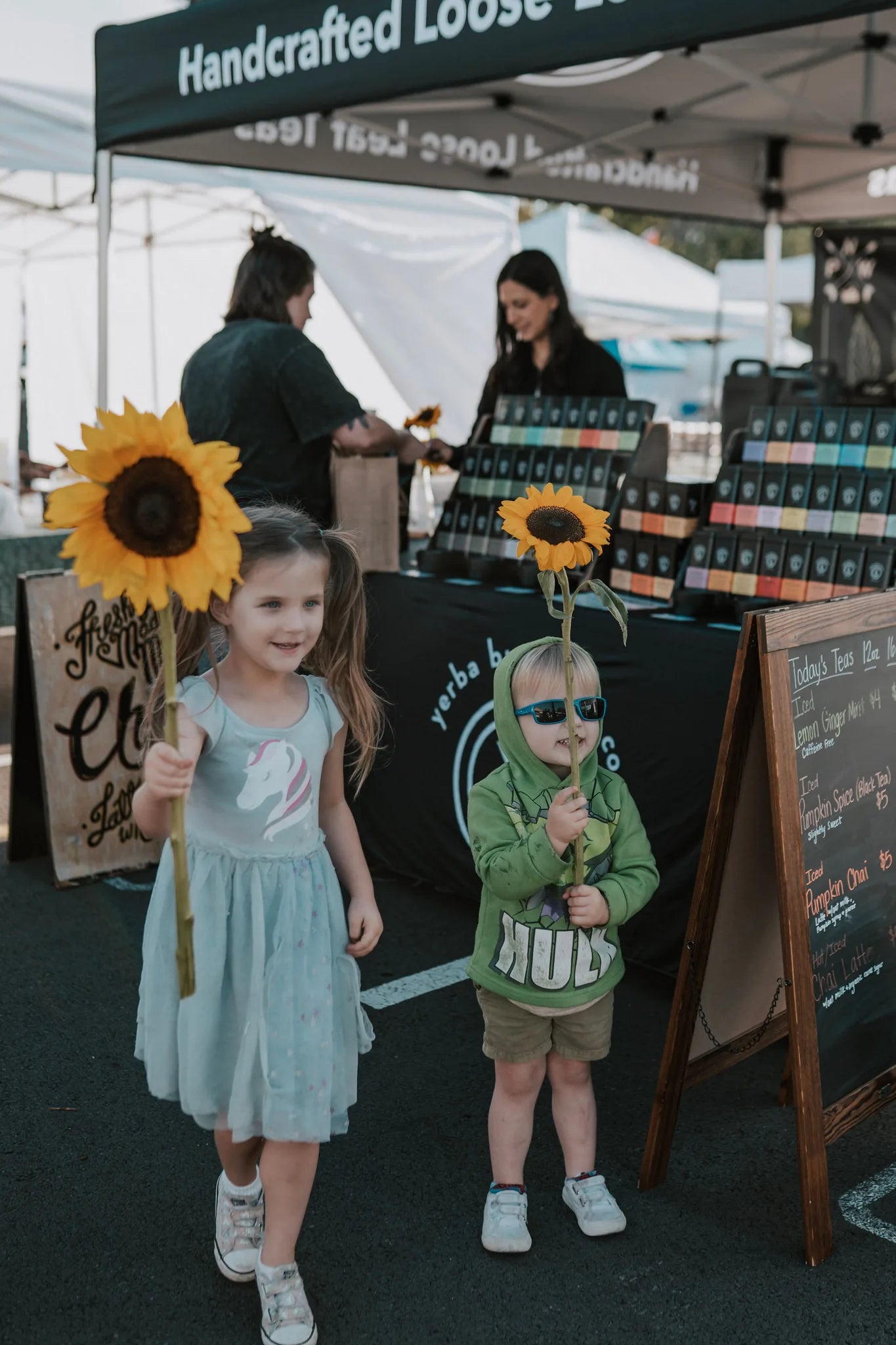 Two young children hold sunflowers at a handcrafted loose leaf tea booth with vendors and colorful tea tin displays visible behind them, Yerba Buena Tea Company.