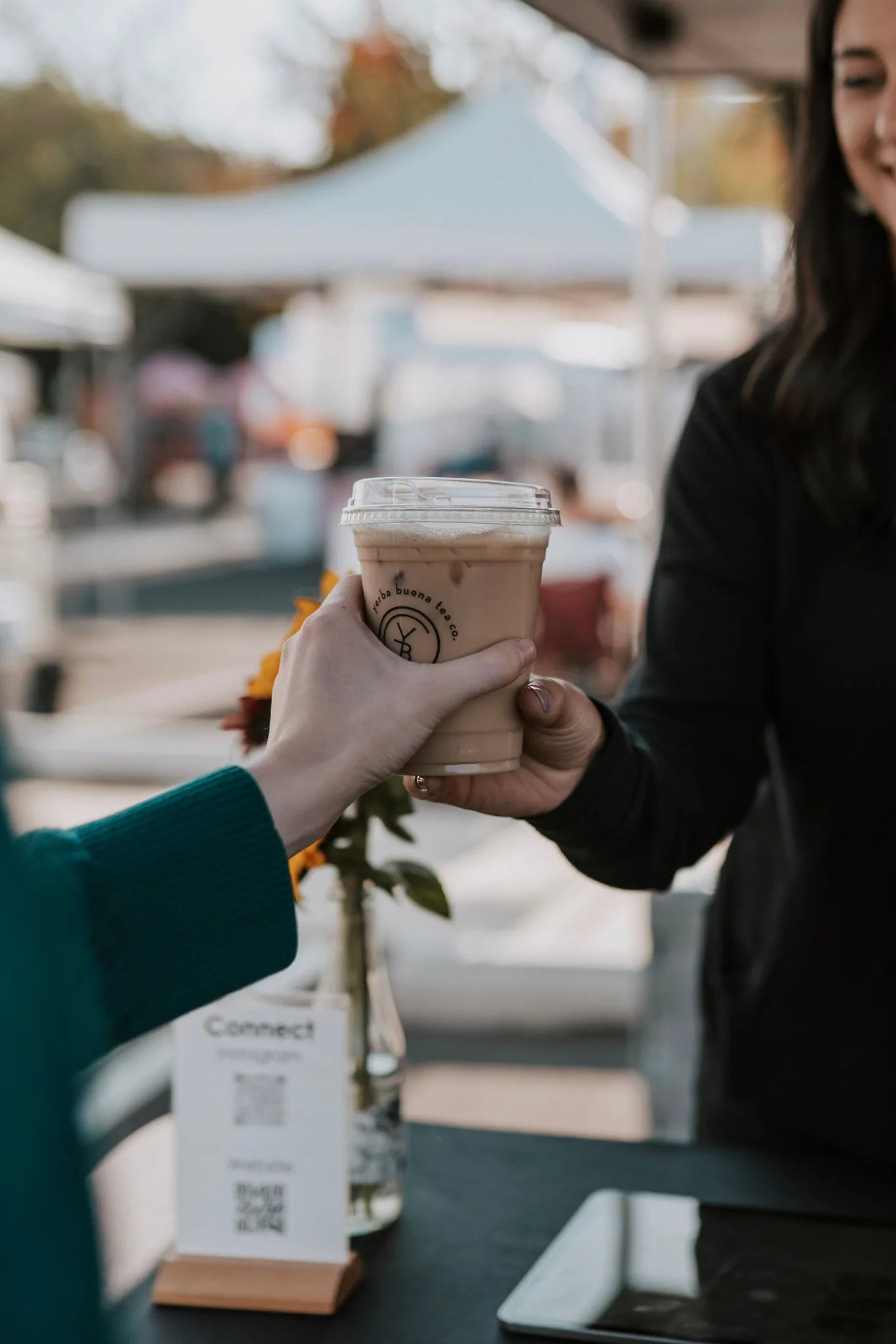 Customer receiving a branded tea beverage at a farmers market booth with white tent canopy and blurred vendor background, Yerba Buena Tea Company.