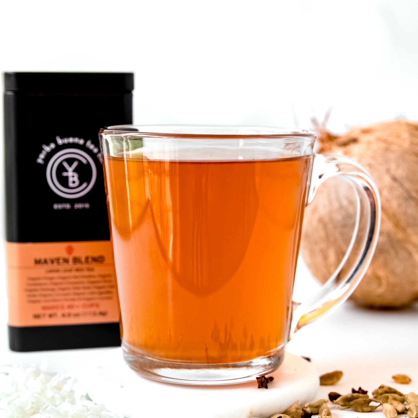 A clear glass cup filled with warm amber Maven Blend tea, placed beside a whole coconut and scattered coconut flakes, with a Yerba Buena Tea Co. tin in the background.