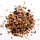 Loose leaf herbal tea blend with dried grass, wood chips, and dark red flower petals scattered on white background, by Yerba Buena Tea Company.