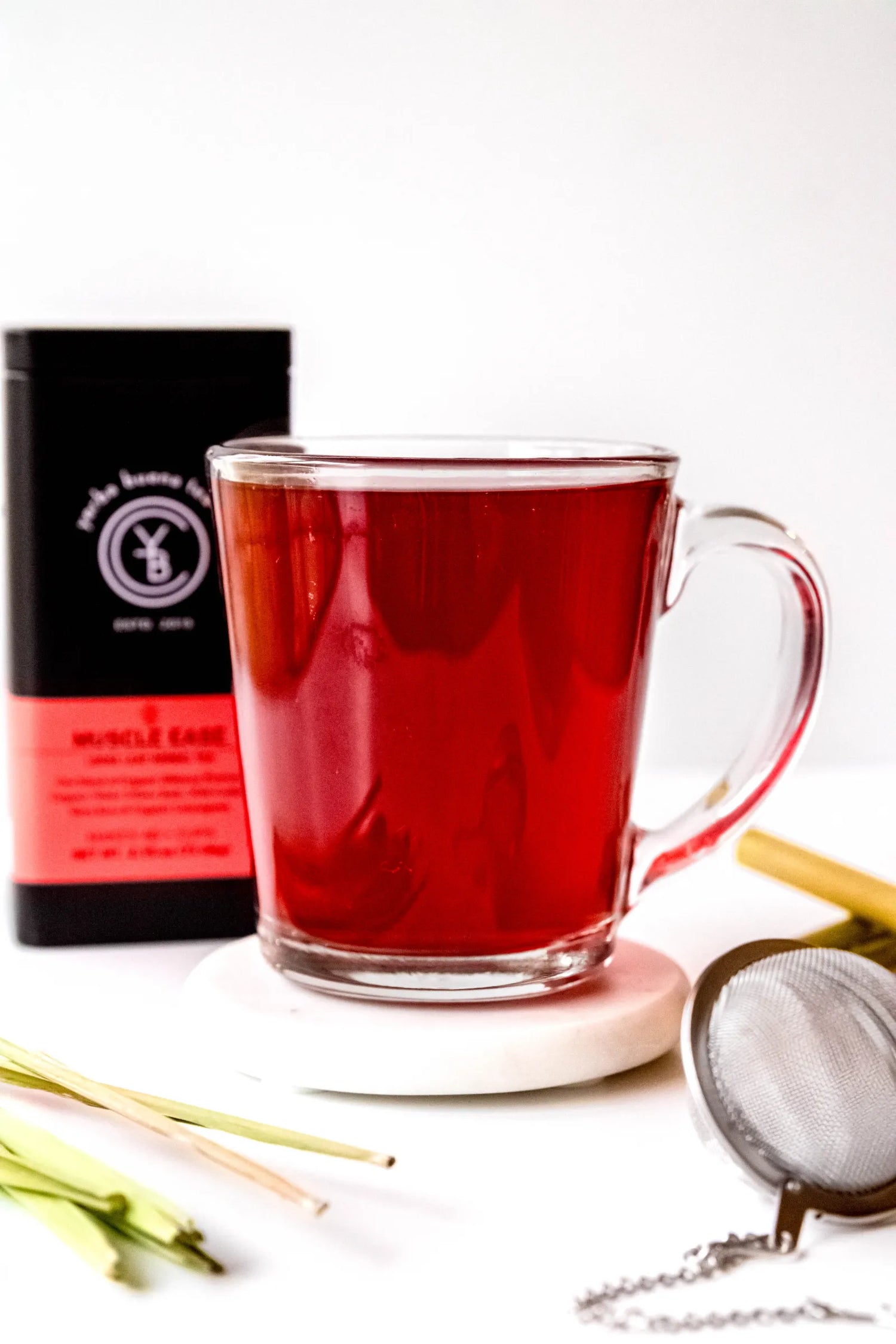 Clear glass mug filled with brewed Muscle Ease organic herbal tea, deep ruby red in color, resting on a white surface beside hibiscus petals and fresh lemongrass. A grounding herbal tea ritual handcrafted by Yerba Buena Tea Co.