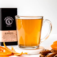 Brewed golden tea in clear glass mug surrounded by orange peel and cinnamon sticks with packaging, by Yerba Buena Tea Company.