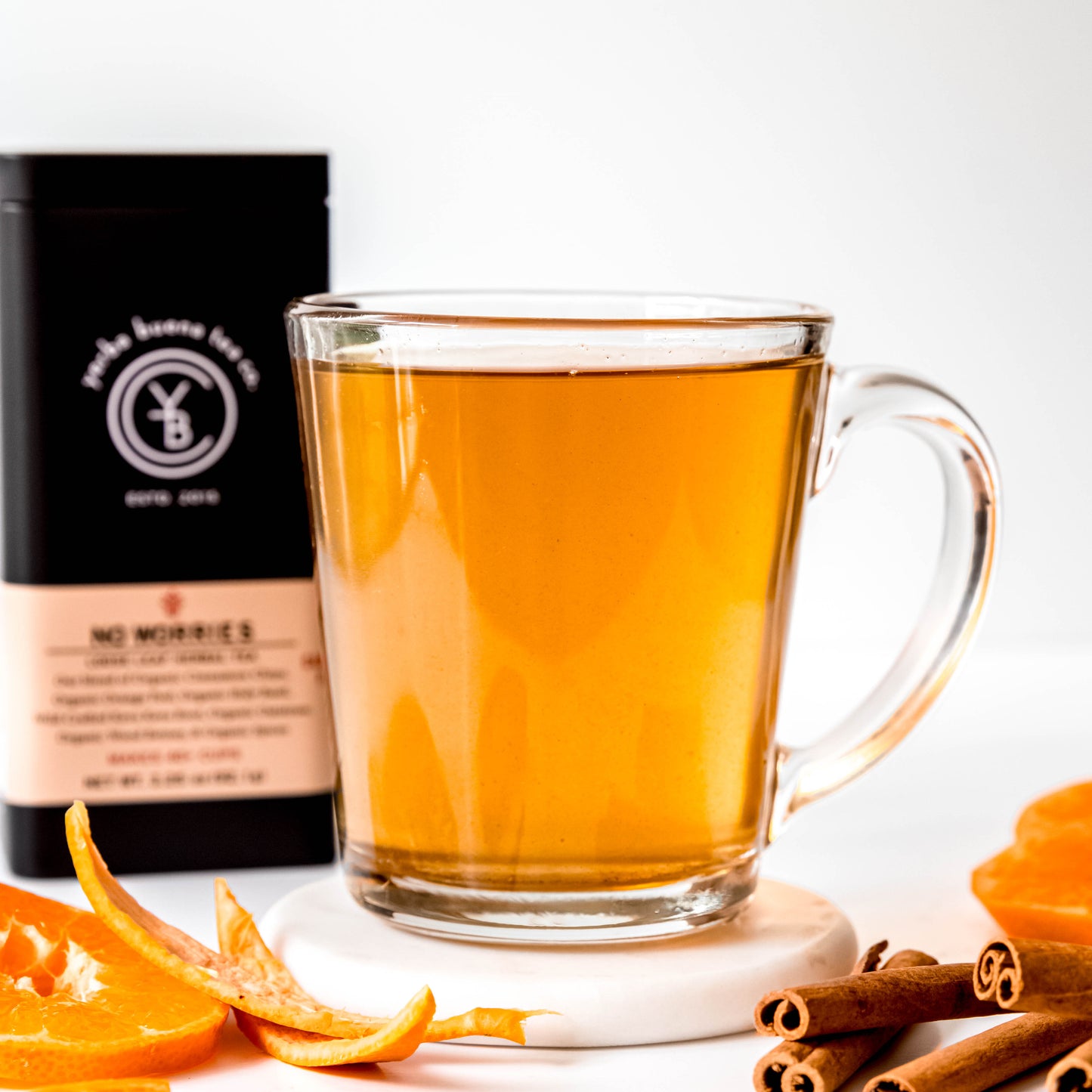 A calming cup of No Worries tea in a clear glass mug, with the Yerba Buena Tea Co. No Worries tea tin in the background. The golden hue of the brewed herbal tea is complemented by fresh orange slices and cinnamon sticks placed beside the cup, creating a serene and inviting setting.