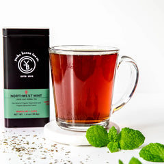 Northwest Mint herbal tea in black canister with green label next to brewed red tea in glass mug and fresh mint leaves, by Yerba Buena Tea Company.