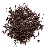 Curled dark brown oolong tea leaves scattered loosely on white background, by Yerba Buena Tea Company.