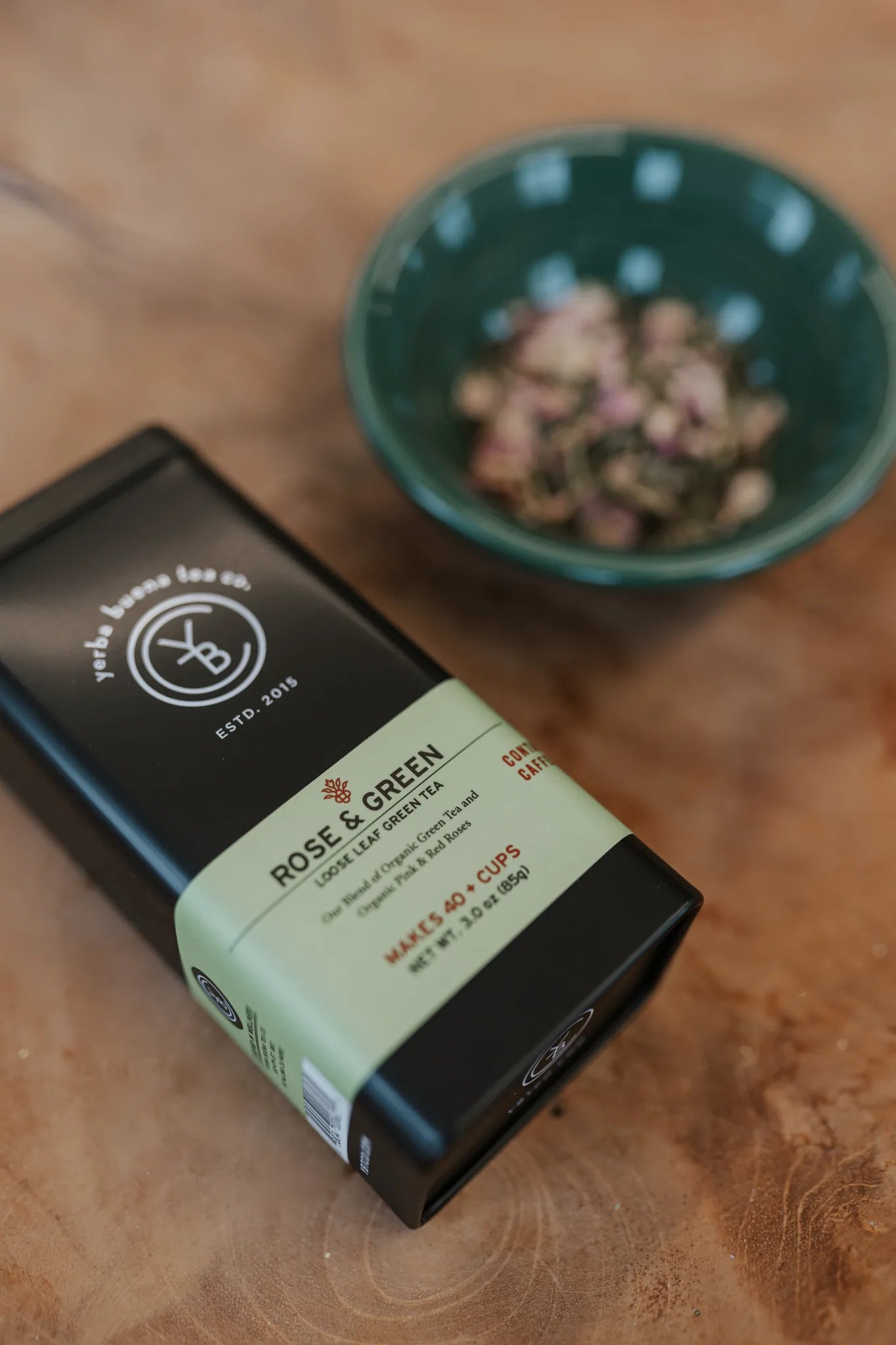Rose & Green tea in a black canister with cream label beside a teal ceramic bowl filled with loose leaf tea on wood surface