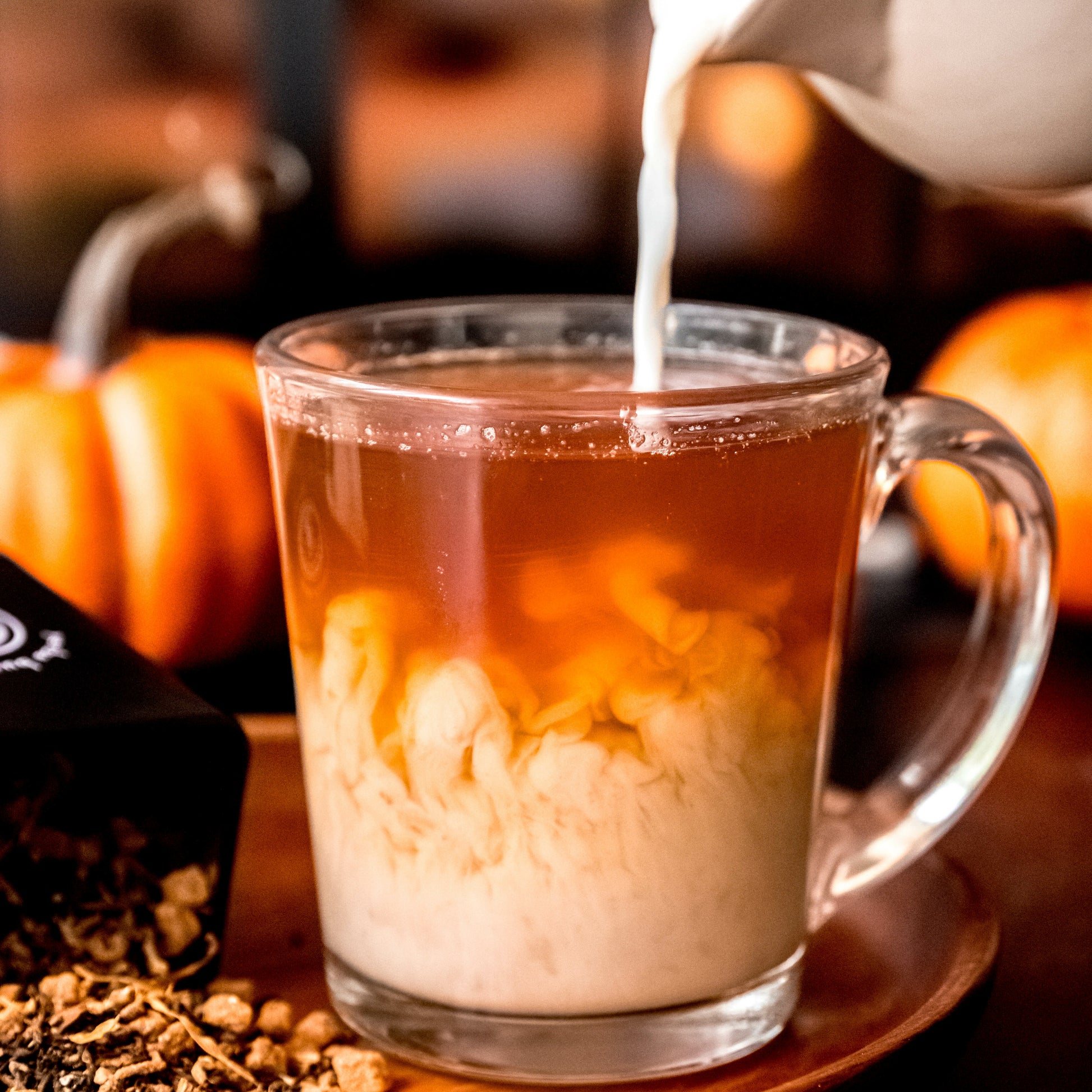 A clear glass cup filled with Pumpkin Spice tea as milk is poured in, creating a swirling effect, with pumpkins in the background and a Yerba Buena Tea Co. tin beside it.