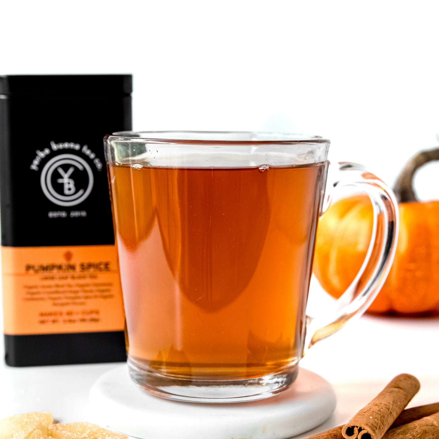 A clear glass cup filled with warm amber Pumpkin Spice tea, placed beside cinnamon sticks and a pumpkin, with a Yerba Buena Tea Co. tin in the background.