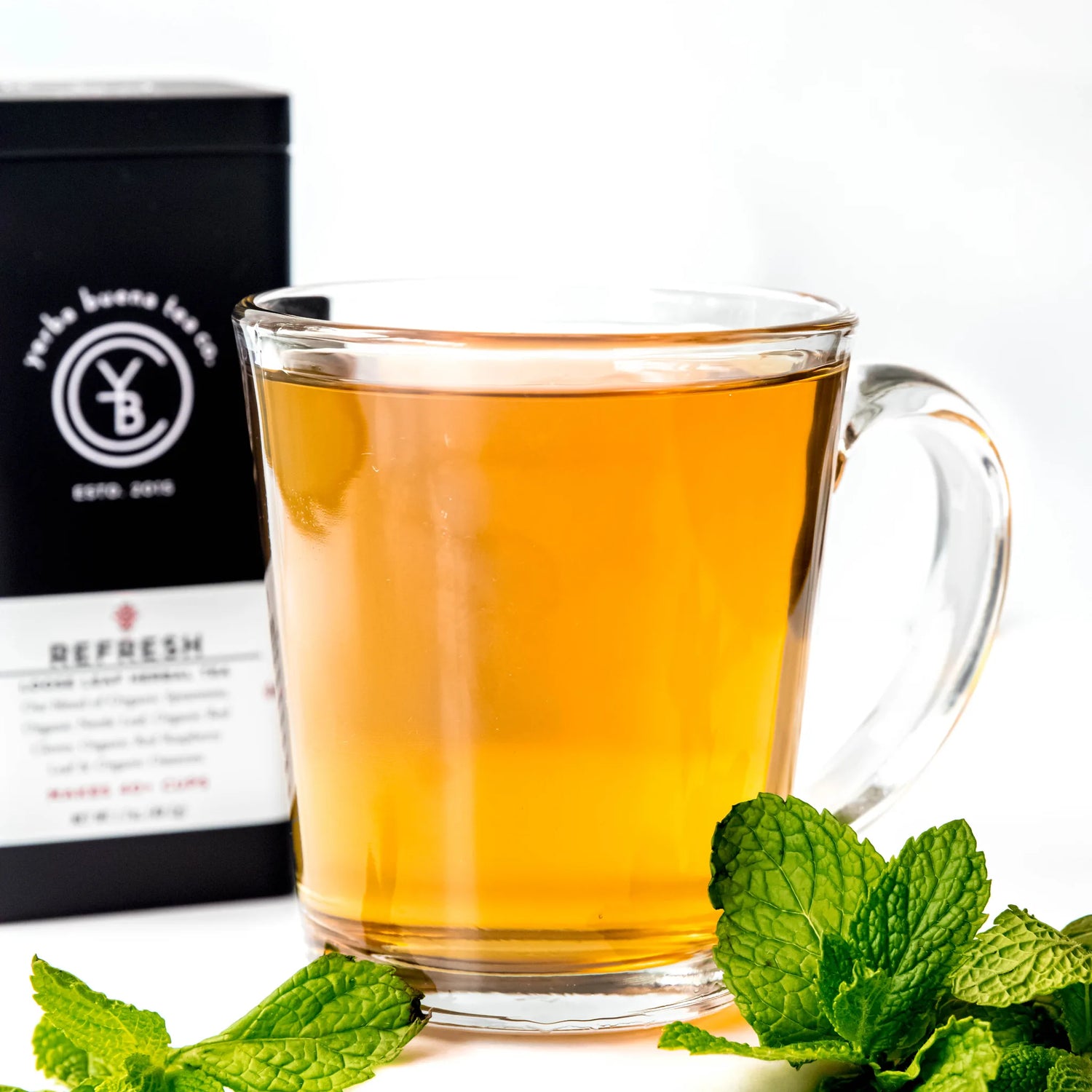 Brewed golden herbal tea in clear glass mug with fresh mint leaves and black packaging, by Yerba Buena Tea Company.