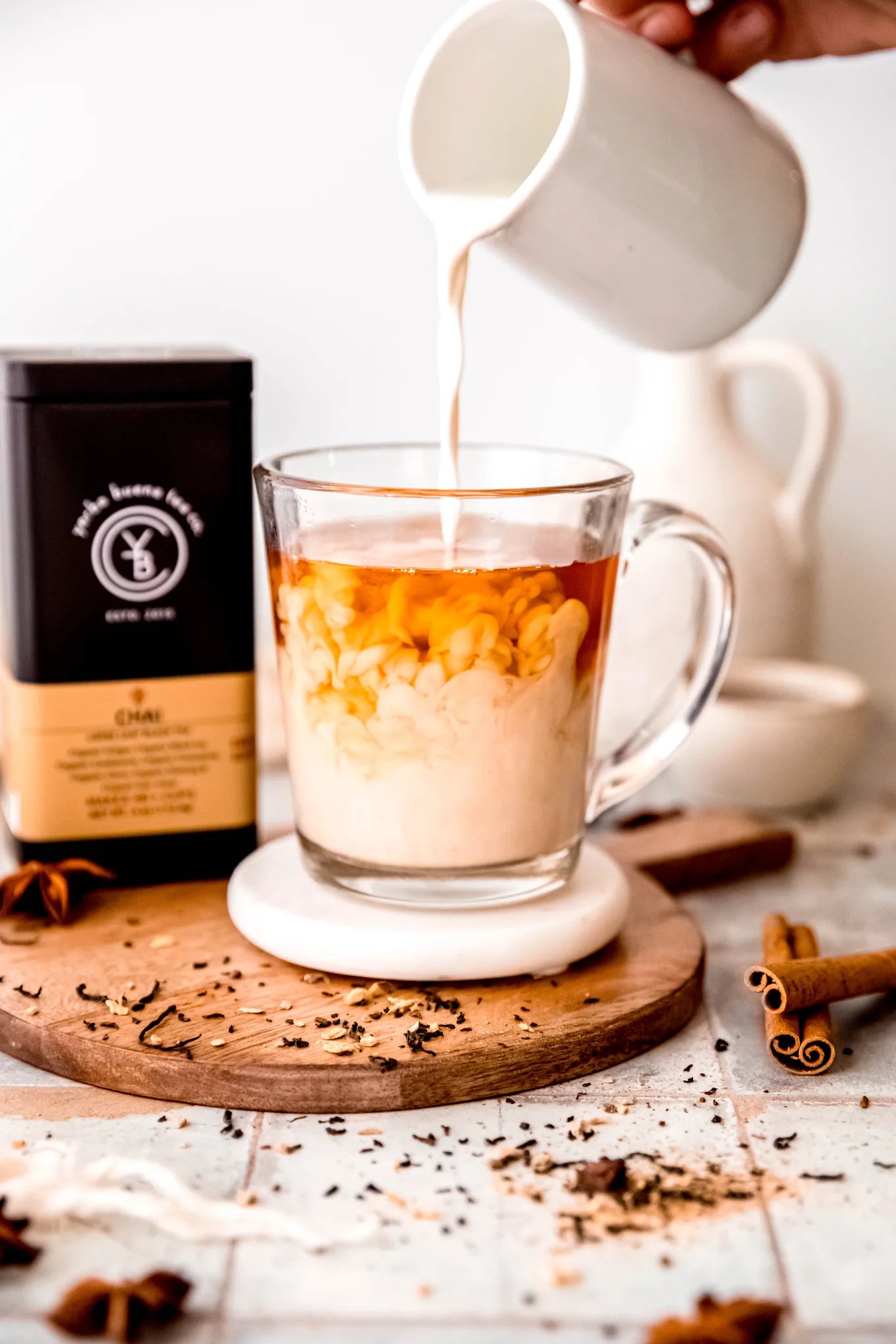 Creamy chai latte being poured from a ceramic pitcher into a glass mug, with loose tea and cinnamon sticks scattered on a wooden board, from Yerba Buena Tea Company.