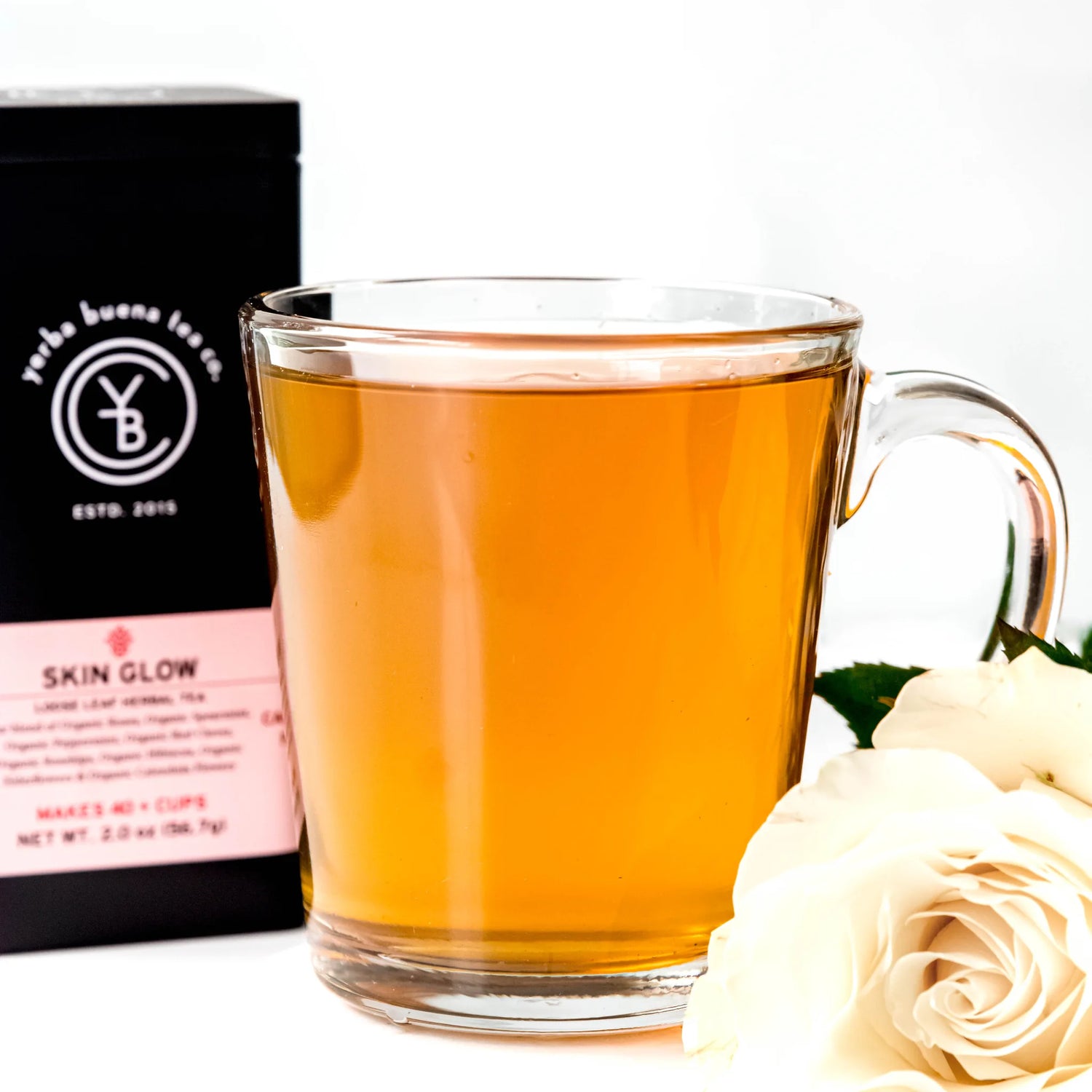 Brewed golden herbal tea in a clear glass mug beside Skin Glow packaging and fresh roses, by Yerba Buena Tea Company.