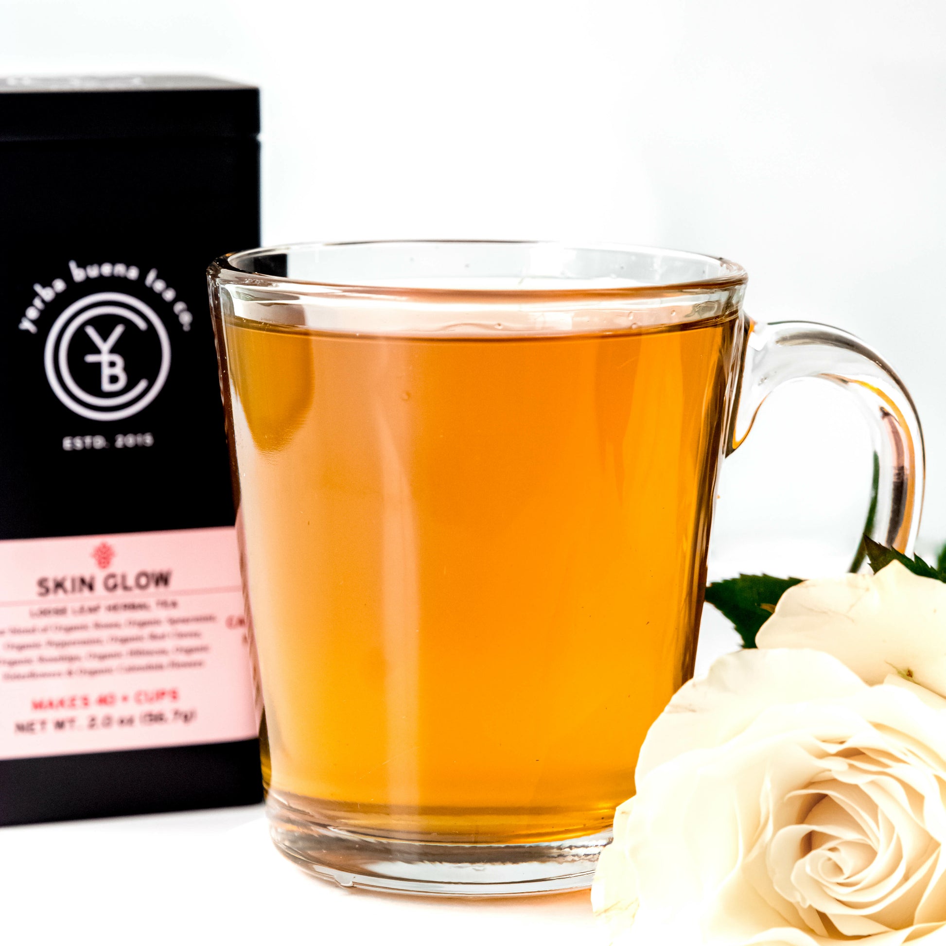 A clear glass cup filled with golden Skin Glow tea, placed beside a white rose, with a Yerba Buena Tea Co. tin in the background.