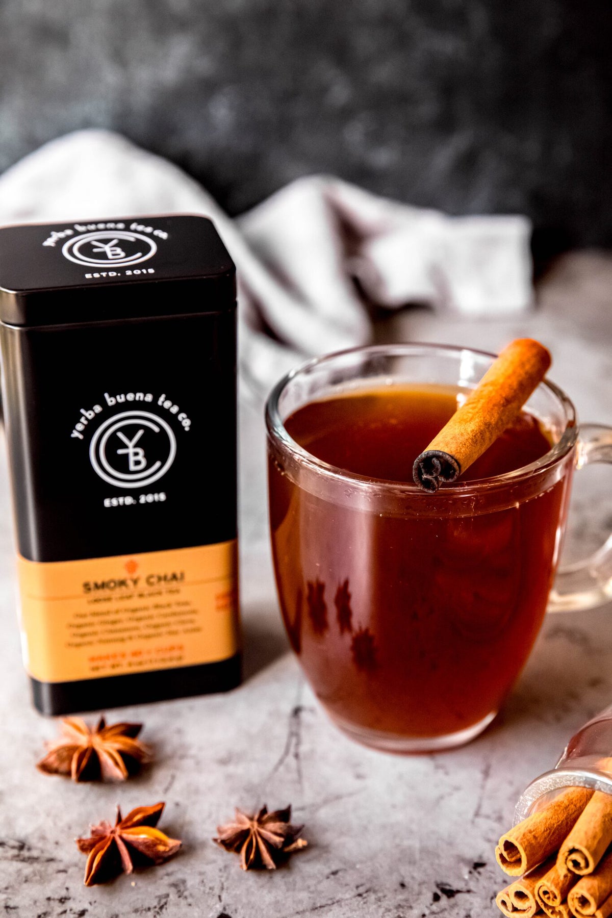 A mug of Smoky Chai tea with a cinnamon stick on a stone counter, beside the Yerba Buena Tea Co. Smoky Chai tin and whole star anise