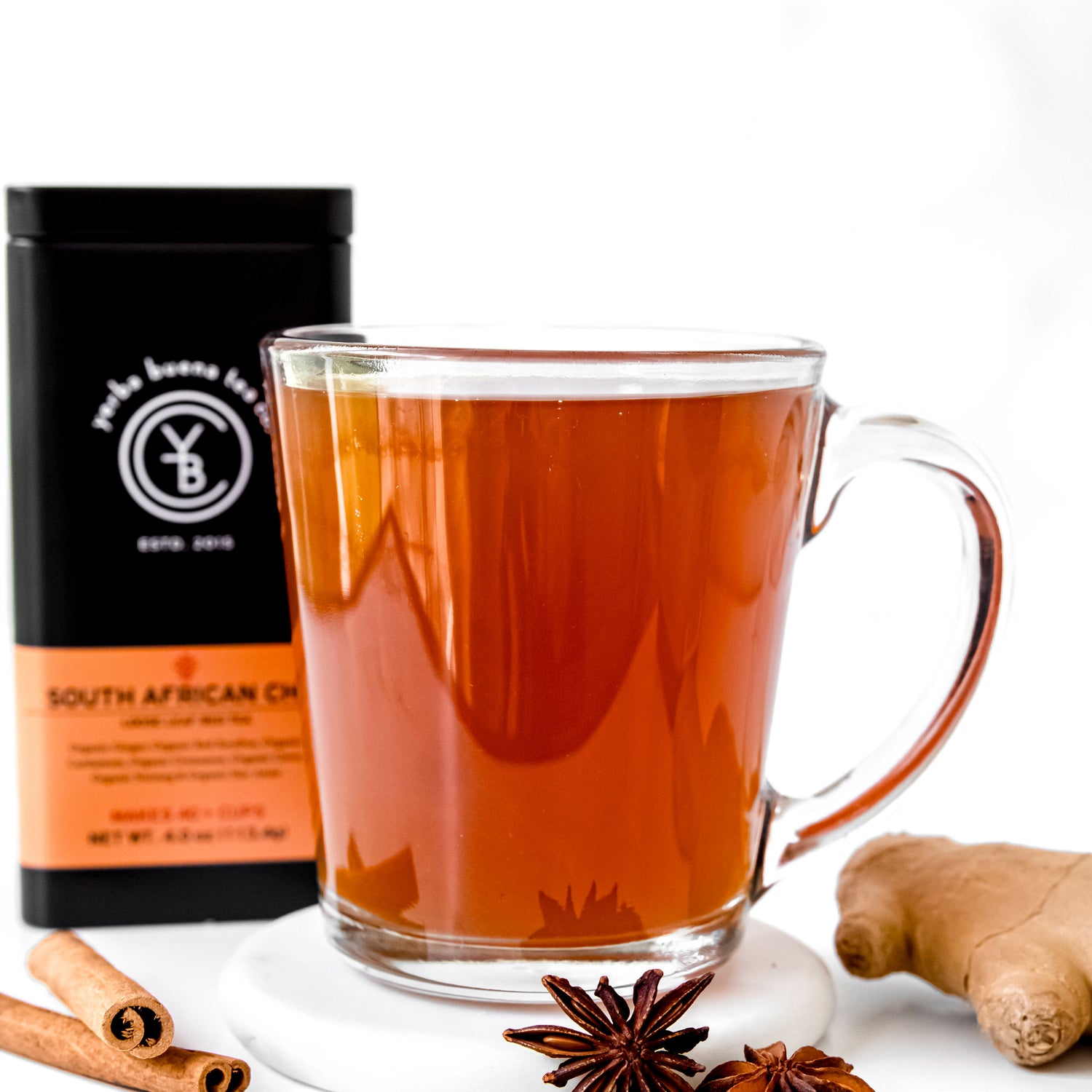 A clear glass cup filled with deep amber South African Chai, placed beside star anise, cinnamon sticks, and ginger root, with a Yerba Buena Tea Co. tin in the background.