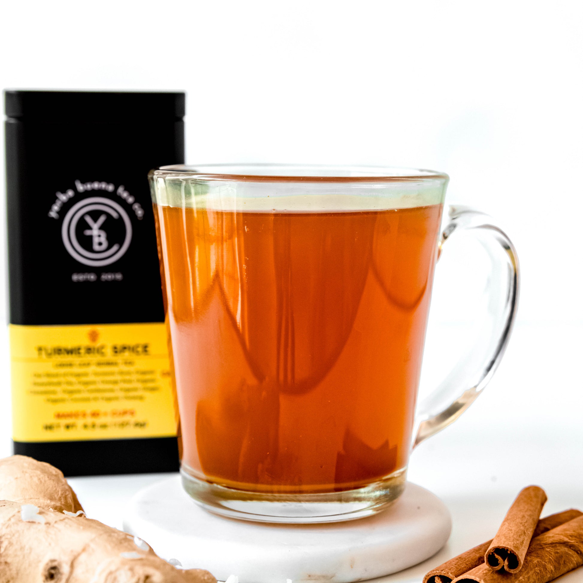 A warm cup of Turmeric Spice tea in a clear glass mug, with the Yerba Buena Tea Co. Turmeric Spice tea tin in the background, showcasing the golden-brown hue of the brewed herbal tea. Fresh ginger and cinnamon sticks are placed beside the cup, enhancing the cozy and inviting setting.