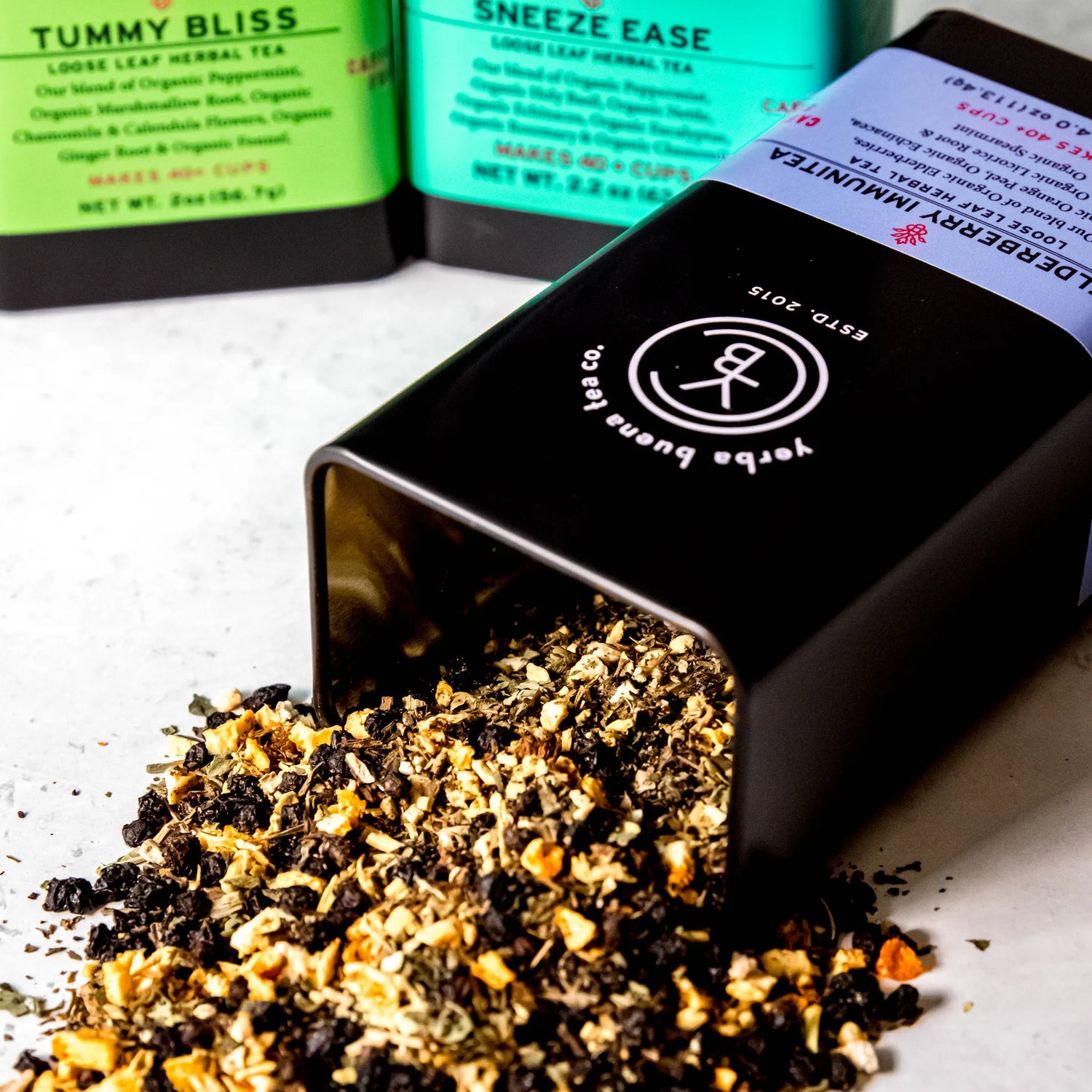 Wellness Feel Better Box Set gift featuring three loose leaf herbal teas: Tummy Bliss, Sneeze Ease, and a third blend, with tea leaves spilling from an open black pouch, by Yerba Buena Tea Company.
