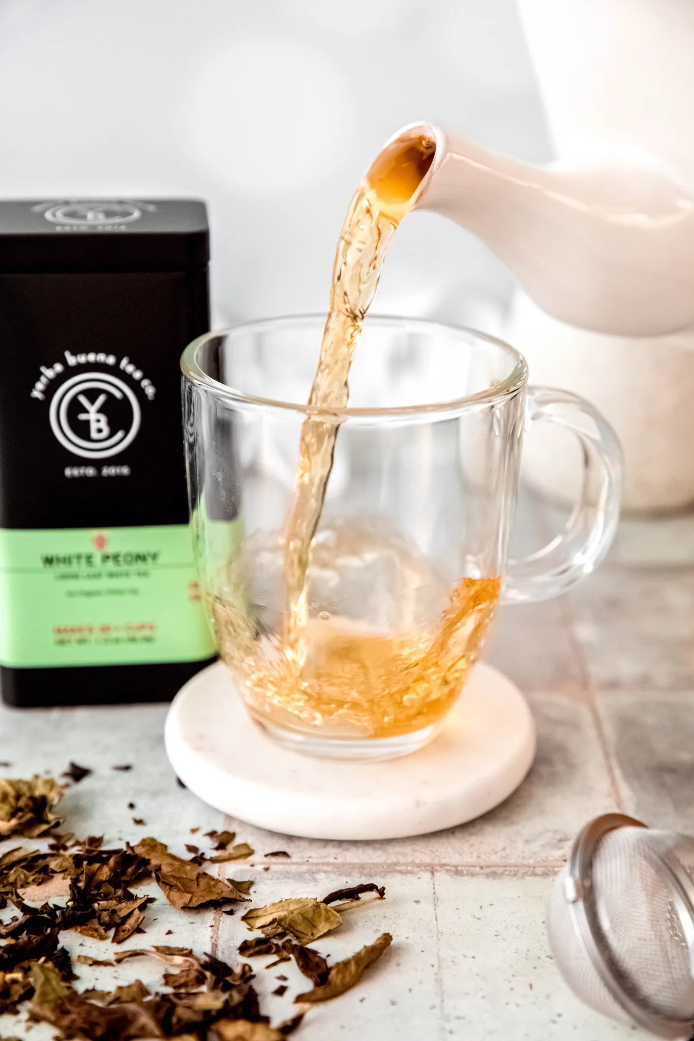 White Peony tea steaming into a clear glass mug from a white ceramic teapot, with loose leaf tea scattered on a light marble surface, by Yerba Buena Tea Company.