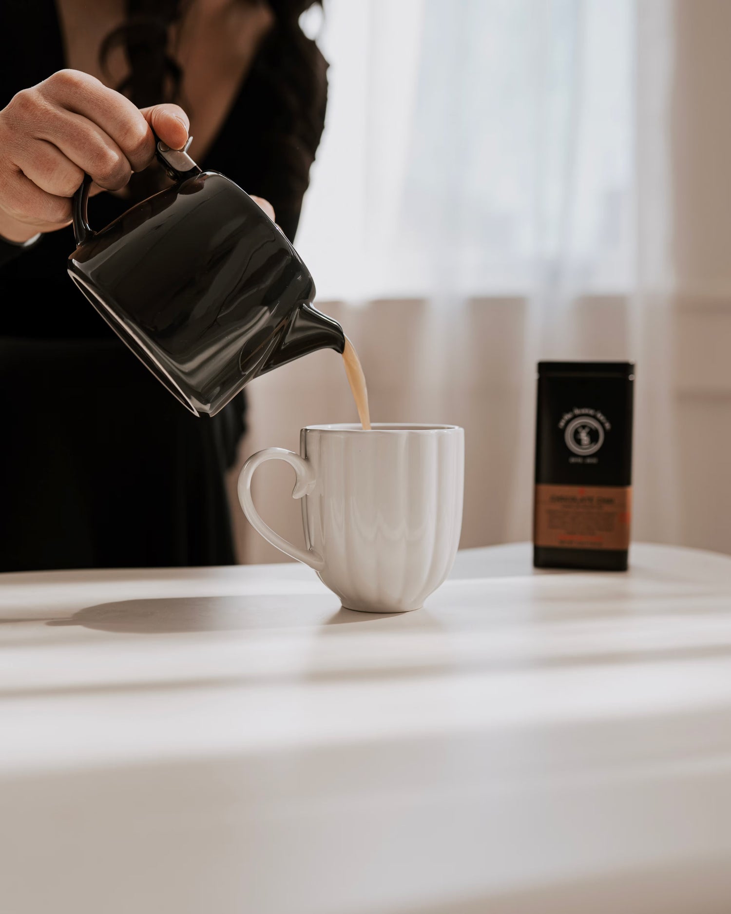 The owner, Hannah, pours a creamy tea latte from a black teapot into a white mug, with a Yerba Buena Tea Co. tin nearby, creating a cozy scene. This moment captures the comforting ritual of a perfect after-dinner treat.