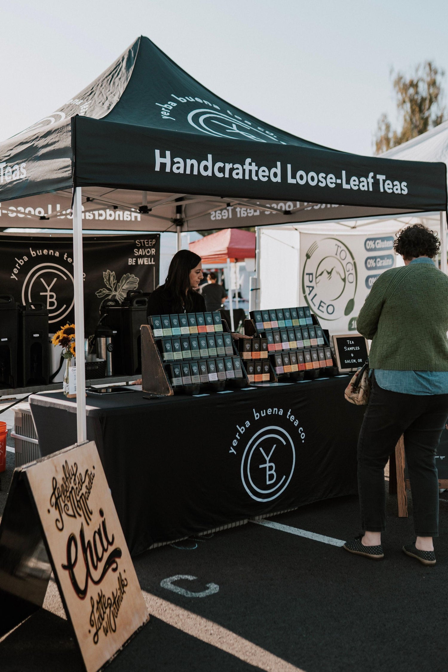 Yerba Buena Tea Co. market booth at an Oregon farmers market displaying organic loose leaf tea tins and hot samples.